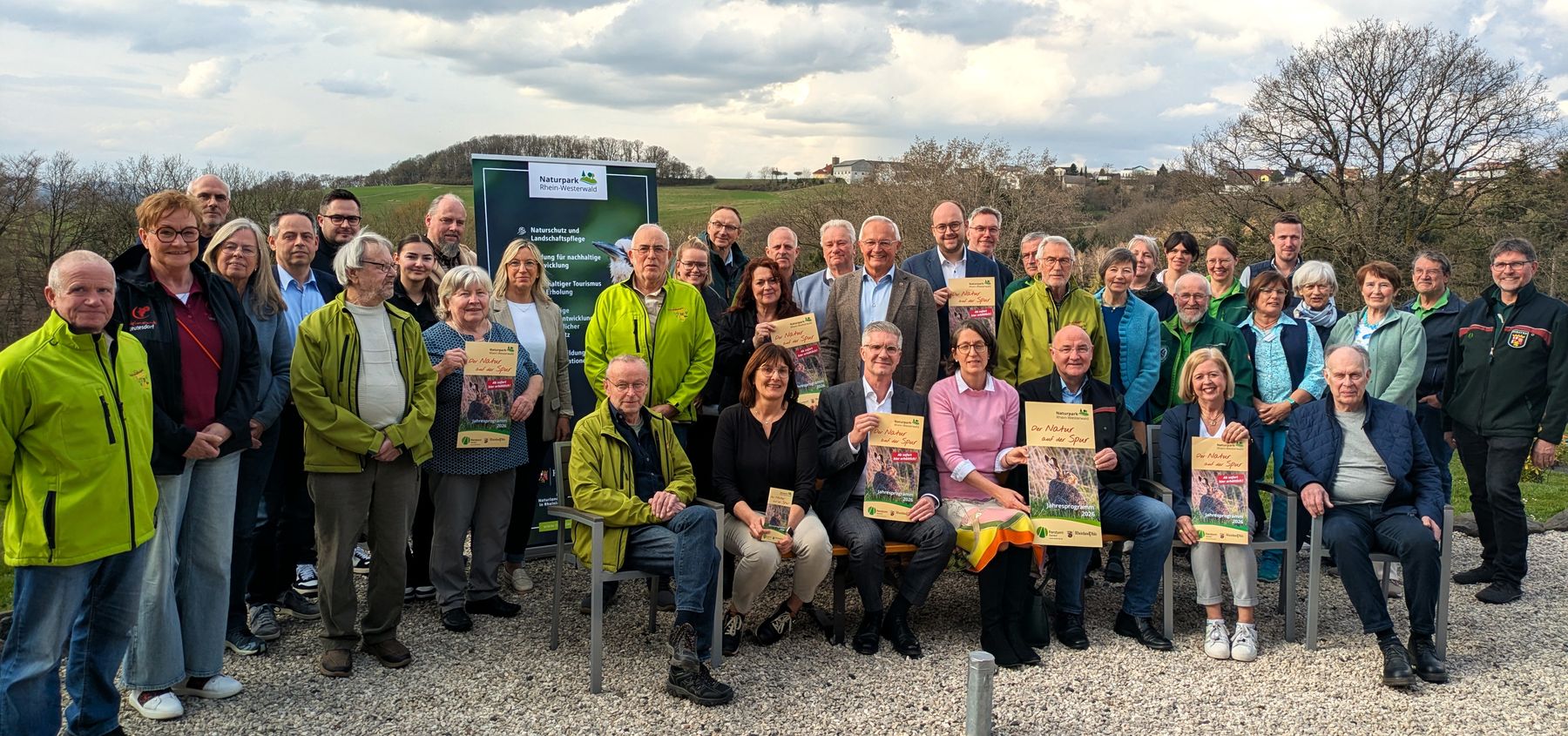 Eine große Gruppe von Menschen, von denen einige Urkunden in der Hand halten, posiert im Freien vor einem grünen Schild, mit Bäumen und einem bewölkten Himmel im Hintergrund. Viele tragen grüne Jacken und scheinen zu feiern oder einer Veranstaltung beizuwohnen.