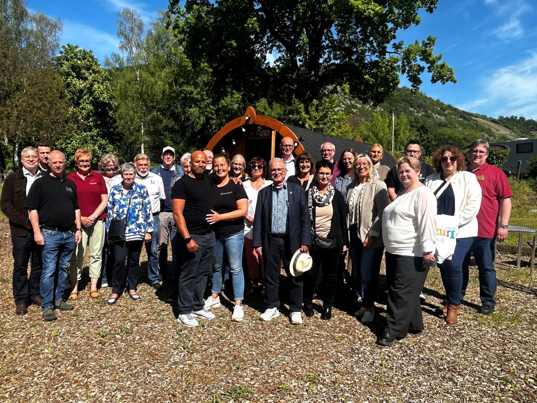 A group of about 25 adults stands outdoors on a sunny day, posing and smiling for a photo in front of a small wooden structure with trees and hills in the background.