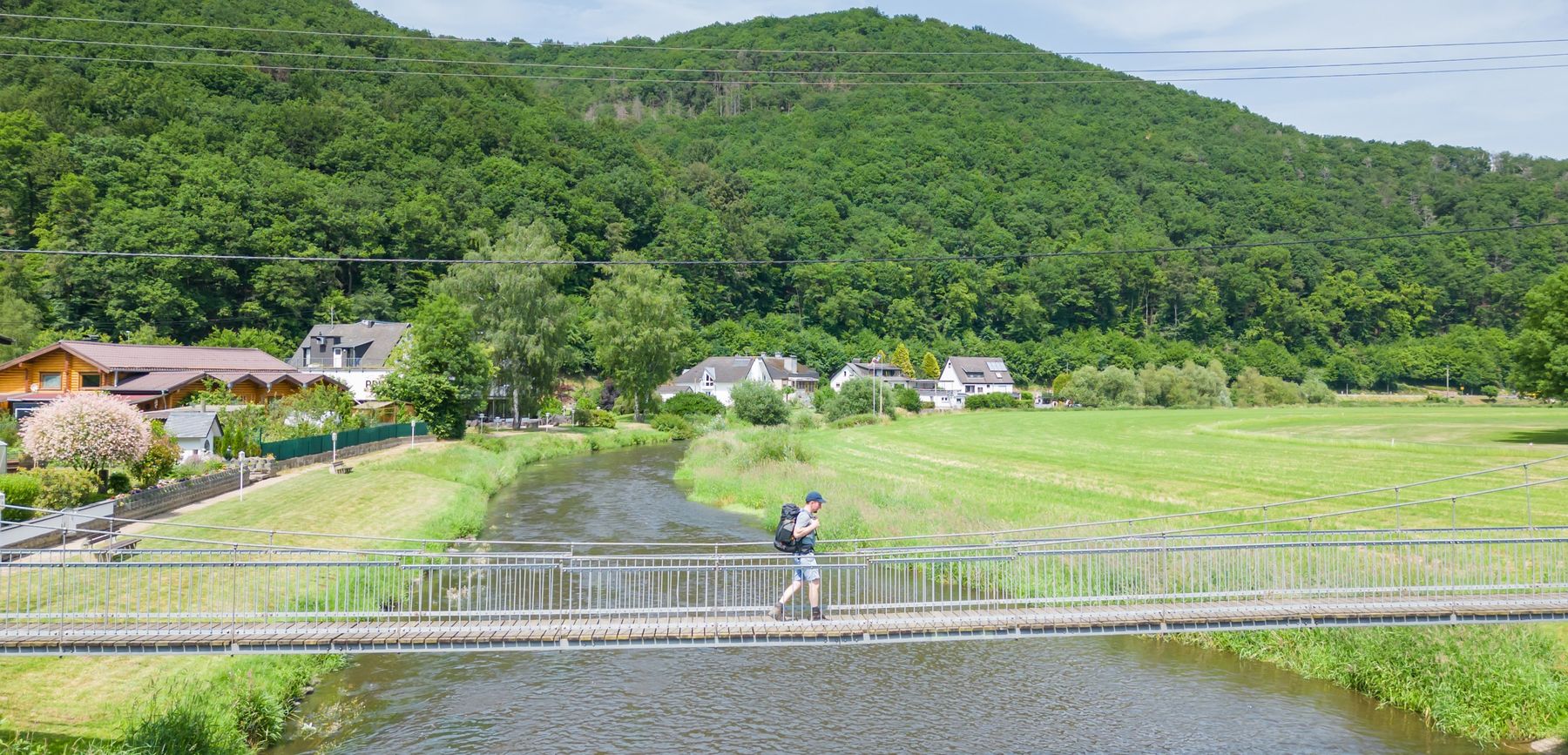 A person with a backpack walks across a narrow suspension bridge over a river, with green fields, houses, and a forested hill in the background under a clear sky.
