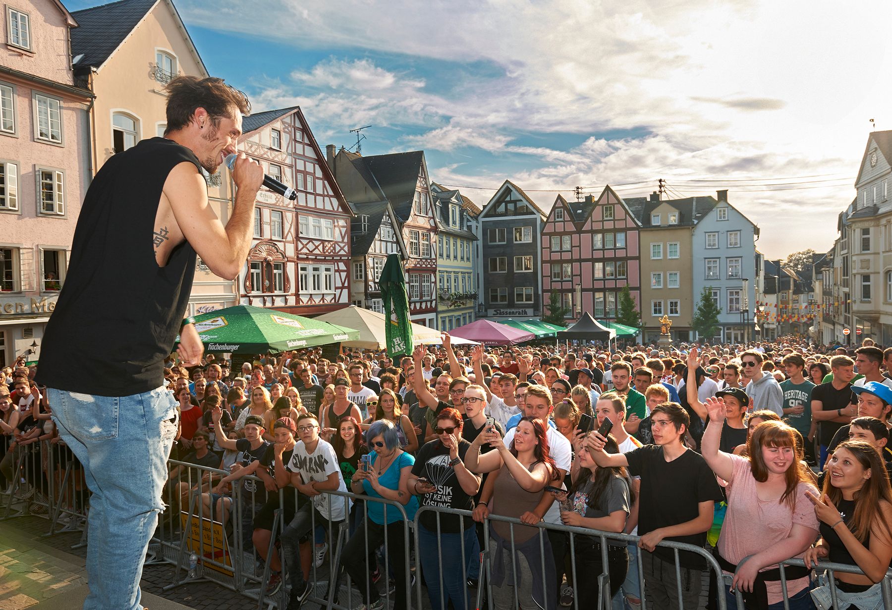 Ein Sänger tritt an einem sonnigen Tag auf einer Freiluftbühne vor einem begeisterten Publikum auf, im Hintergrund sind farbenfrohe historische Gebäude und Zelte zu sehen.